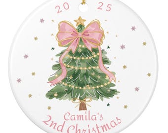 May include: A round white ceramic ornament featuring a green Christmas tree illustration with a large pink bow on top, gold star, and string lights. The text 20 25 appears at the top, and the text Camila's 2nd Christmas is printed in pink at the bottom.