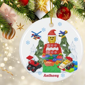May include: A white ceramic Christmas ornament featuring a Lego minifigure wearing a red and white Santa hat and sweater. The minifigure is holding a pile of colorful Lego bricks. The ornament also includes a red truck, a white car, and a helicopter. The ornament is personalized with the name "Anthony" and the year "2024".