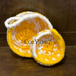 May include: Two handmade, woven baskets in yellow and white. The larger basket has a handle. The text "Cozy YarnZ" is visible on the baskets. The baskets are made of a soft, textured material.