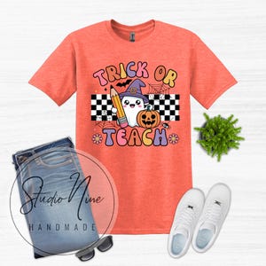 May include: A coral-colored short-sleeve t-shirt featuring a graphic of a ghost wearing a purple witch hat, holding a pencil next to a pumpkin. The text reads TRICK OR TEACH, surrounded by a black and white checkerboard pattern and spiderwebs.