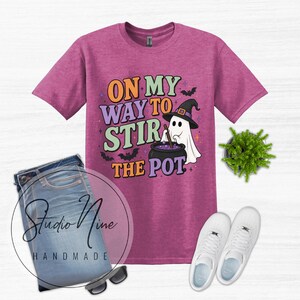 May include: A heathered magenta t-shirt featuring a graphic of a ghost wearing a witch hat and stirring a cauldron. The text reads ON MY WAY TO STIR THE POT, accompanied by small bat and star illustrations. The shirt is paired with blue jeans.