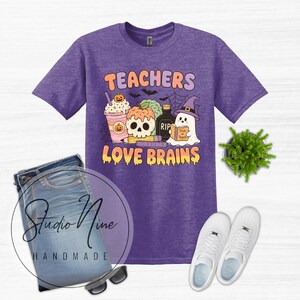 May include: A heather purple short-sleeve t-shirt featuring a graphic print with the text "TEACHERS LOVE BRAINS". The design includes a pumpkin-topped drink, a skull, a bottle labeled "RIP", a ghost holding a book, a pencil, and a ruler.