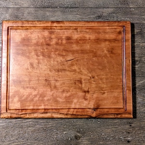 May include: A rectangular wooden cutting board with a juice groove. The board is made of light brown wood with a natural grain pattern. The edges are slightly rounded, and the board has a smooth, polished finish. The cutting board is a kitchen essential.