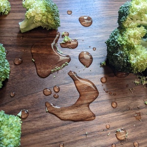 May include: Overhead shot of fresh broccoli florets and water droplets on a dark brown wooden surface. The broccoli is a vibrant green, and the water droplets reflect light, creating a contrast with the wood grain.