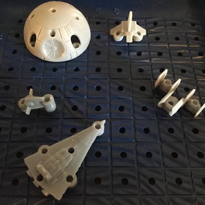 May include: A close-up of a Star Wars game board with five plastic game pieces. The pieces are a Death Star, a TIE fighter, a TIE Interceptor, a X-Wing, and a Y-Wing. The board is blue and has a grid pattern.
