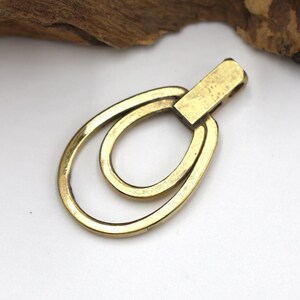 May include: A gold-colored metal pendant with a rectangular top and two connected oval loops. The pendant is set against a white background with a piece of brown wood in the upper left corner.