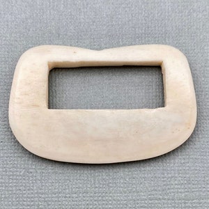 May include: A white bone toggle with a rectangular opening in the center. The toggle is smooth and has a slightly rounded shape.