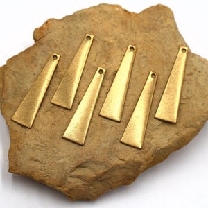 May include: Seven brass-colored, elongated triangle charms with small holes at the top are arranged on a rough, tan stone. The charms have a brushed metal texture and a slight taper from the base to the top.