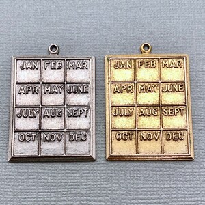 May include: Two metal charms, one silver and one gold, with a calendar design. Each charm has a grid with the months of the year, January through December, printed on it.