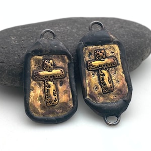 May include: Two black and gold cross charms with a textured surface. The charms have a loop at the top for attaching to jewelry.