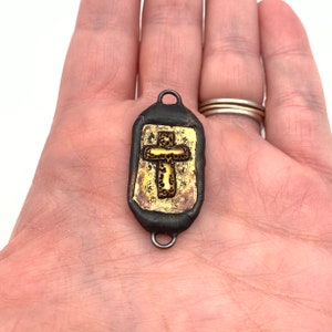 May include: A black and gold pendant with a cross design. The pendant is made of metal and has a textured surface.