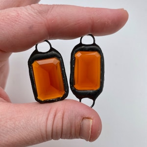 May include: Two orange glass gems set in black metal frames. The gems are rectangular with rounded corners and have a slightly cloudy appearance.