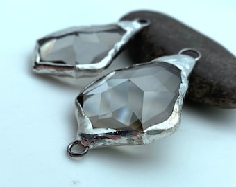 May include: Two clear glass teardrop-shaped pendants with silver metal frames. The pendants have a faceted design and are hanging from a silver metal loop.