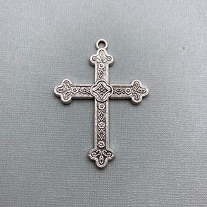 May include: A silver cross pendant with an ornate design. The cross has a detailed floral pattern and a small, raised center piece.