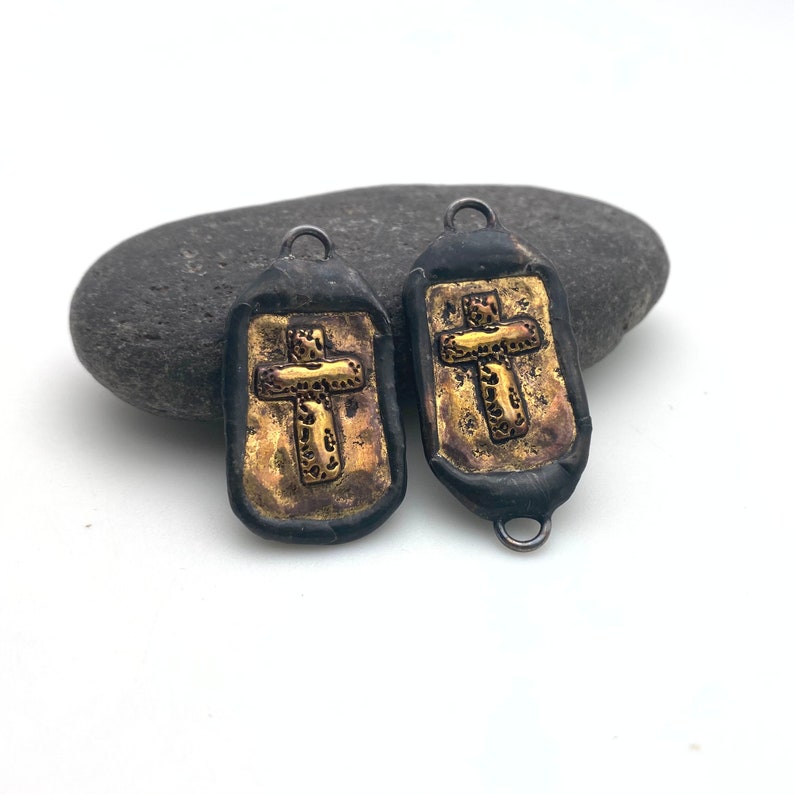 May include: Two black and gold cross charms with a textured surface. The charms have a loop at the top for attaching to jewelry.