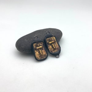 May include: Two black and gold cross charms with a textured surface. The charms are hanging from a rock.