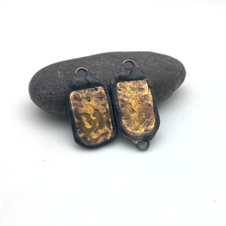 May include: Two black metal pendants with gold leaf accents. The pendants are rectangular and have a slightly textured surface.