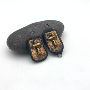 May include: Two black metal charms with gold cross designs. The charms are shaped like a rectangle with rounded corners. The crosses are raised and have a textured surface.