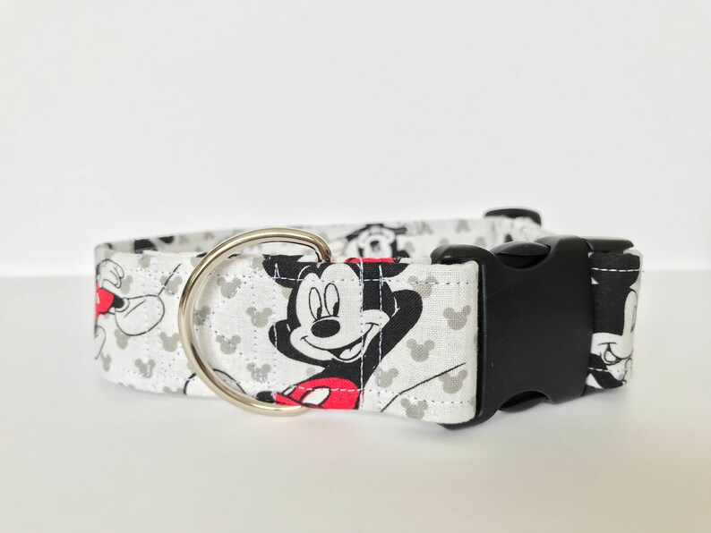 K&ouml;nnte beinhalten: Wei&szlig;er Hundehalsband mit einer schwarzen Schnalle und einem silbernen Ring. Das Halsband hat ein sich wiederholendes Muster aus Mickey Mouse-K&ouml;pfen in Schwarzwei&szlig; mit roten Akzenten.