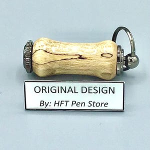 Pode incluir: Uma caneta artesanal com um corpo de madeira clara e padr&otilde;es de gr&atilde;o escuros. Possui detalhes em metal e um porta-chaves. O texto "ORIGINAL DESIGN By: HFT Pen Store" est&aacute; em uma etiqueta branca.
