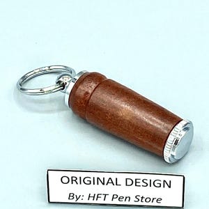 Pode incluir: Um porta-canetas cil&iacute;ndrico de madeira castanha com detalhes em metal prateado e um porta-chaves. O porta-canetas tem um acabamento polido. O texto "ORIGINAL DESIGN By: HFT Pen Store" &eacute; vis&iacute;vel numa etiqueta branca.