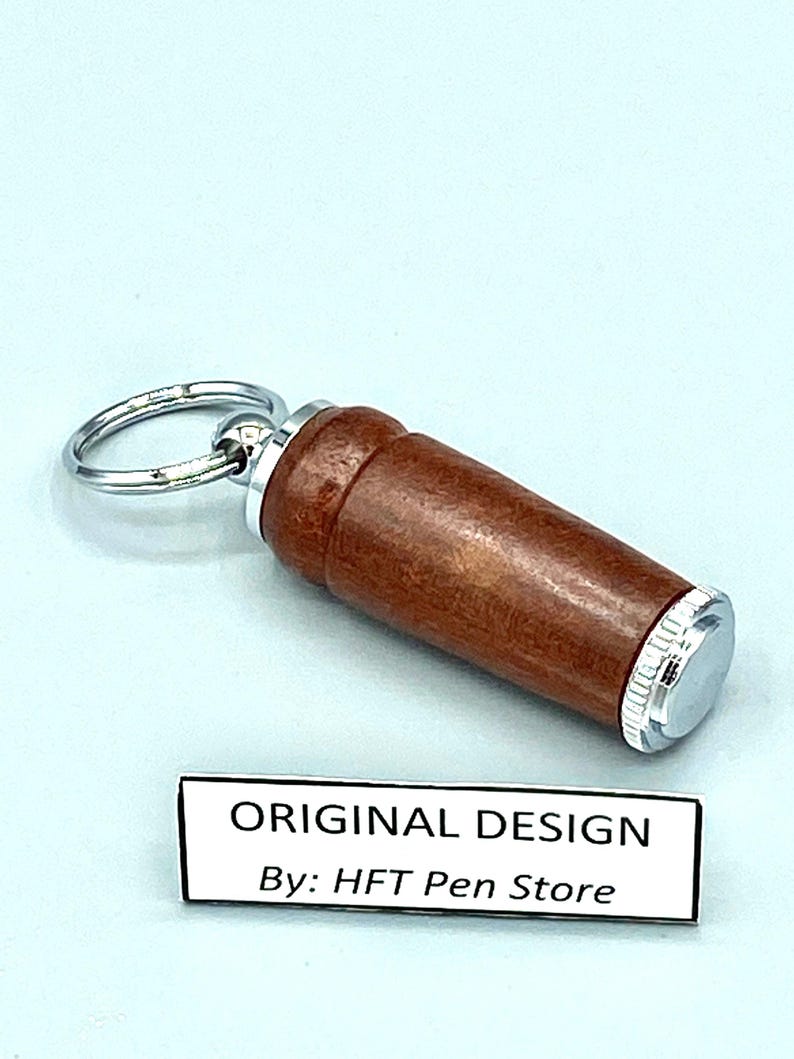 Pode incluir: Um porta-canetas cil&iacute;ndrico de madeira castanha com detalhes em metal prateado e um porta-chaves. O porta-canetas tem um acabamento polido. O texto "ORIGINAL DESIGN By: HFT Pen Store" &eacute; vis&iacute;vel numa etiqueta branca.