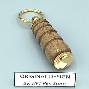 Pode incluir: Um porta-chaves de madeira com um anel e detalhes dourados. O porta-chaves tem um design cil&iacute;ndrico com uma s&eacute;rie de an&eacute;is esculpidos. O texto "ORIGINAL DESIGN By: HFT Pen Store" &eacute; exibido em um r&oacute;tulo branco.
