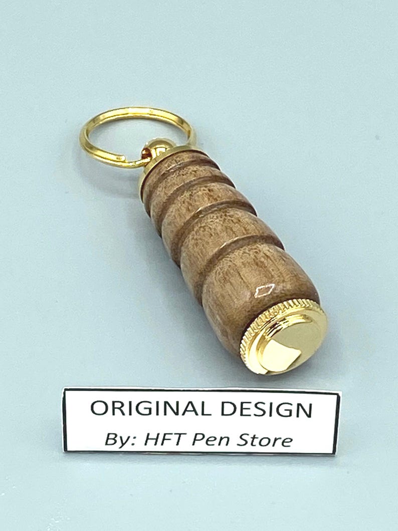 Pode incluir: Um porta-chaves de madeira com um anel e detalhes dourados. O porta-chaves tem um design cil&iacute;ndrico com uma s&eacute;rie de an&eacute;is esculpidos. O texto "ORIGINAL DESIGN By: HFT Pen Store" &eacute; exibido em um r&oacute;tulo branco.
