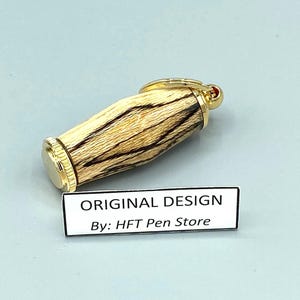 Pode incluir: Uma caneta com tampa e detalhes dourados, e um corpo de madeira com listras marrons escuras e bege. A caneta tem um pequeno detalhe vermelho na parte superior. O texto "ORIGINAL DESIGN By: HFT Pen Store" est&aacute; em uma etiqueta branca.