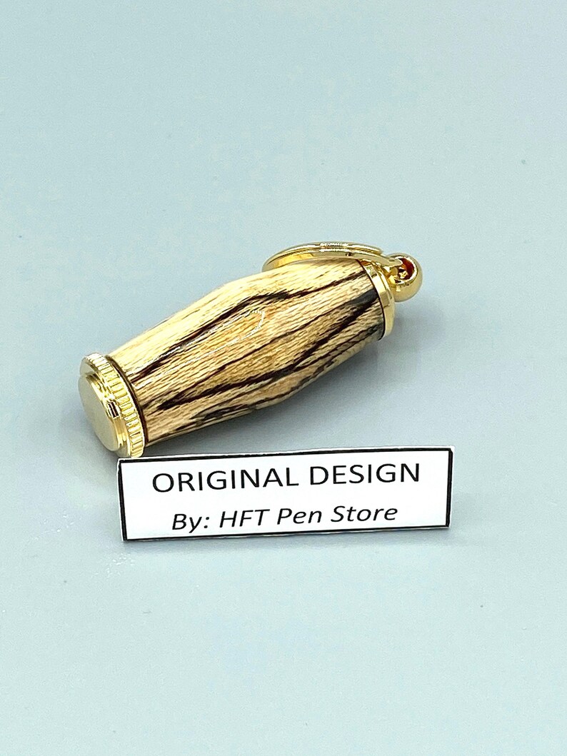 Pode incluir: Uma caneta com tampa e detalhes dourados, e um corpo de madeira com listras marrons escuras e bege. A caneta tem um pequeno detalhe vermelho na parte superior. O texto "ORIGINAL DESIGN By: HFT Pen Store" est&aacute; em uma etiqueta branca.