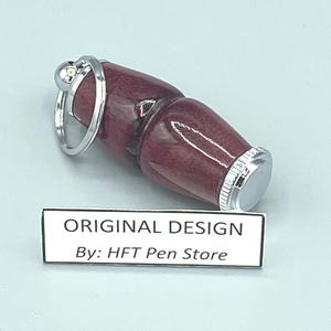Pode incluir: Uma caneta bord&ocirc; com um anel e detalhes em metal prateado. A caneta tem um acabamento liso e polido e uma ponta de metal prateado. O texto "ORIGINAL DESIGN By: HFT Pen Store" &eacute; exibido em um r&oacute;tulo branco.