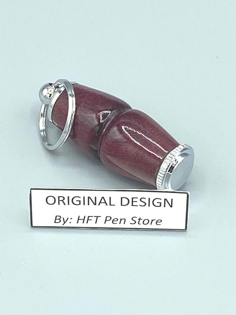Pode incluir: Uma caneta bord&ocirc; com um anel e detalhes em metal prateado. A caneta tem um acabamento liso e polido e uma ponta de metal prateado. O texto "ORIGINAL DESIGN By: HFT Pen Store" &eacute; exibido em um r&oacute;tulo branco.