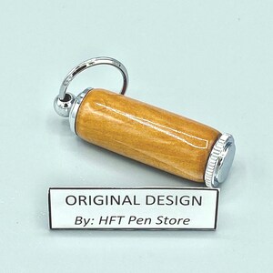 Pode incluir: Um porta-canetas de madeira cil&iacute;ndrico com tampa e chaveiro de metal prateado. O porta-canetas tem um acabamento castanho claro. O texto "ORIGINAL DESIGN By: HFT Pen Store" est&aacute; impresso em uma etiqueta branca.