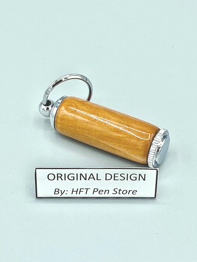 Pode incluir: Um porta-canetas de madeira cil&iacute;ndrico com tampa e chaveiro de metal prateado. O porta-canetas tem um acabamento castanho claro. O texto "ORIGINAL DESIGN By: HFT Pen Store" est&aacute; impresso em uma etiqueta branca.