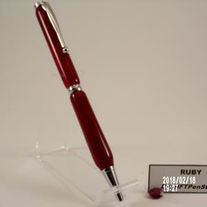 Può includere: Una penna rossa e argentata con un clip sulla parte superiore. La penna è su un supporto in acrilico trasparente. Il testo "RUBY 2018/02/18 19:27 FTPenStore" è su un'etichetta bianca.