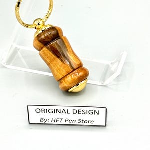 Pode incluir: Um porta-chaves de madeira artesanal com um anel e detalhes dourados. O porta-chaves apresenta um design cil&iacute;ndrico com um padr&atilde;o de gr&atilde;o de madeira natural e um acabamento brilhante. O texto "ORIGINAL DESIGN By: HFT Pen Store" &eacute; exibido em um r&oacute;tulo branco.