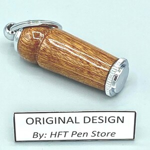 Pode incluir: Um porta-canetas cil&iacute;ndrico de madeira com tampa e chaveiro de metal prateado. O porta-canetas tem um padr&atilde;o de gr&atilde;o de madeira castanho claro. O texto "ORIGINAL DESIGN By: HFT Pen Store" est&aacute; em uma etiqueta branca.