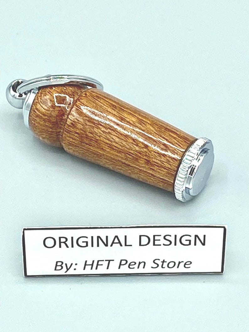 Pode incluir: Um porta-canetas cil&iacute;ndrico de madeira com tampa e chaveiro de metal prateado. O porta-canetas tem um padr&atilde;o de gr&atilde;o de madeira castanho claro. O texto "ORIGINAL DESIGN By: HFT Pen Store" est&aacute; em uma etiqueta branca.