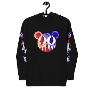 Puede incluir: Sudadera con capucha negra con un gráfico colorido similar a un oso en la parte delantera con un diseño rojo, blanco y azul. Las mangas tienen un diseño repetido de cohete y oso. La sudadera tiene una capucha con cordón y un bolsillo tipo canguro.