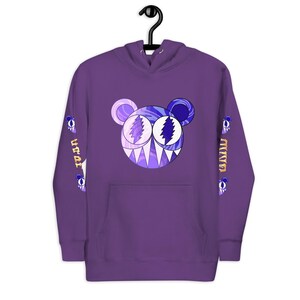 May include: A purple hoodie with a large cartoon bear face on the front. The bear has a white and purple face with blue and purple accents. The sleeves have text and small bear graphics. The hoodie has a front pocket and a hood.