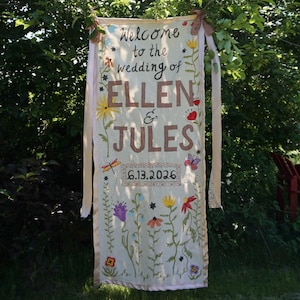 Könnte beinhalten: Ein rechteckiges, cremefarbenes Stoffbanner mit dem handgemalten Text Welcome to the wedding of ELLEN & JULES 6.13.2026. Das Banner ist mit bunten Blumenillustrationen, Schmetterlingen und Libellen verziert und hängt an zwei hellen Bändern.