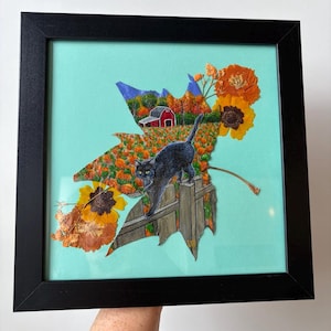 May include: A framed artwork featuring a large dried leaf painted with a scene of a black cat walking on a wooden fence in front of a pumpkin patch and a red barn. The composition is accented with pressed orange flowers on a light blue background.