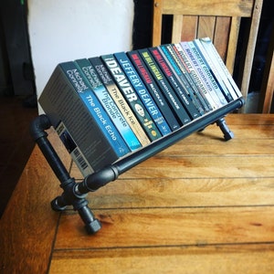 May include: A black metal pipe bookshelf with a variety of books on it. The books are mostly paperback novels with titles like "The Black Ice" and "From the Dead".