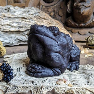 May include: A dark brown frog-shaped candle with detailed texture. The candle sits on a lace doily, with a wooden background and decorative elements. The candle has a wick and is a unique decorative item.