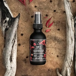 May include: A black spray bottle with a label that reads "IGNITE RITUAL SPRAY". The label features red floral illustrations and text. The bottle is surrounded by driftwood and red leaves on a textured surface.