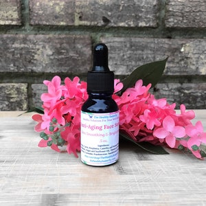 May include: A dark blue glass bottle with a black dropper, labeled "Anti-Aging Face Serum." The bottle is surrounded by pink flowers and green leaves. The label also includes the text "Skin Smoothing & Brightening" and "5 oz."