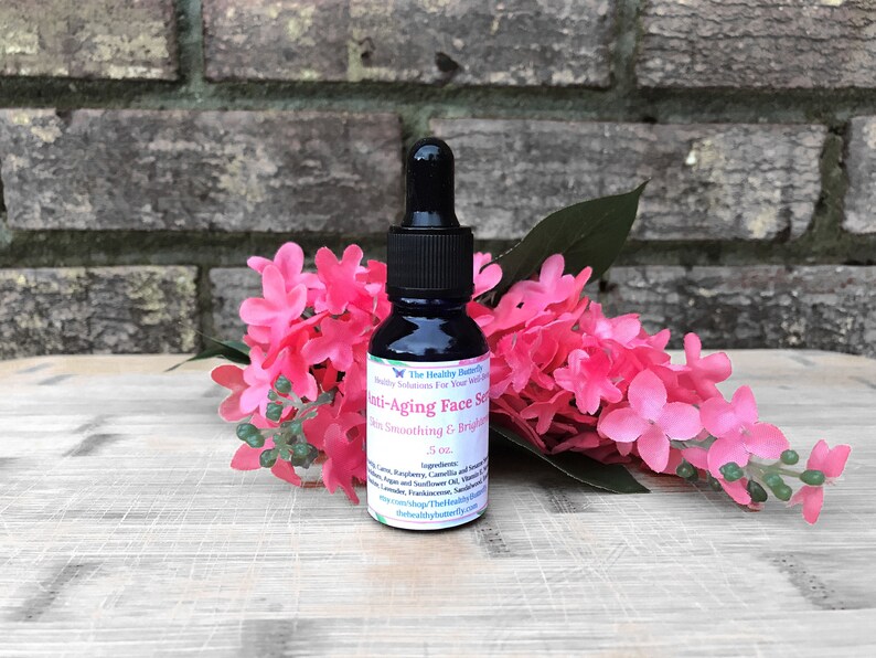 May include: A dark blue glass bottle with a black dropper, labeled "Anti-Aging Face Serum." The bottle is surrounded by pink flowers and green leaves. The label also includes the text "Skin Smoothing & Brightening" and "5 oz."