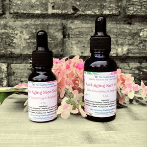 May include: Two dark glass bottles of Anti-Aging Face Serum with dropper applicators. The labels read "Skin Smoothing & Brightening" and list ingredients. One bottle is .5 oz, the other is 1 oz. Pink flowers and greenery are in the background.