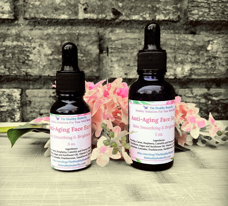 May include: Two dark glass bottles of Anti-Aging Face Serum with dropper applicators. The labels read "Skin Smoothing & Brightening" and list ingredients. One bottle is .5 oz, the other is 1 oz. Pink flowers and greenery are in the background.