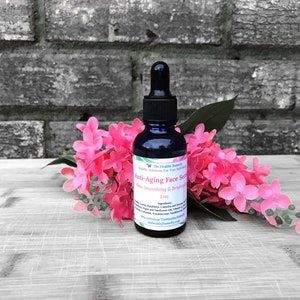 May include: A dark blue glass bottle of anti-aging face serum with a black dropper, set against a backdrop of pink flowers and a gray brick wall. The label reads "Anti-Aging Face Serum, Skin Smoothing & Brightening."